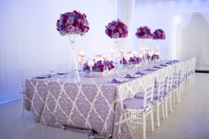 Table set with white tablecloth and purple flowers with white chairs around it