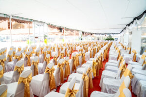 Temporary Air Conditioner indoor tent for outdoor event in the day. Inside Tents has red carpet and a lot of chair covered by white cloth and golden ribbon on it.